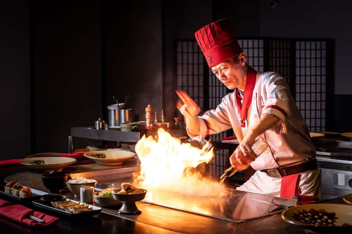 Ein Koch bereitet ein Flambieren in einer Küche mit traditionellem japanischen Hintergrund vor.