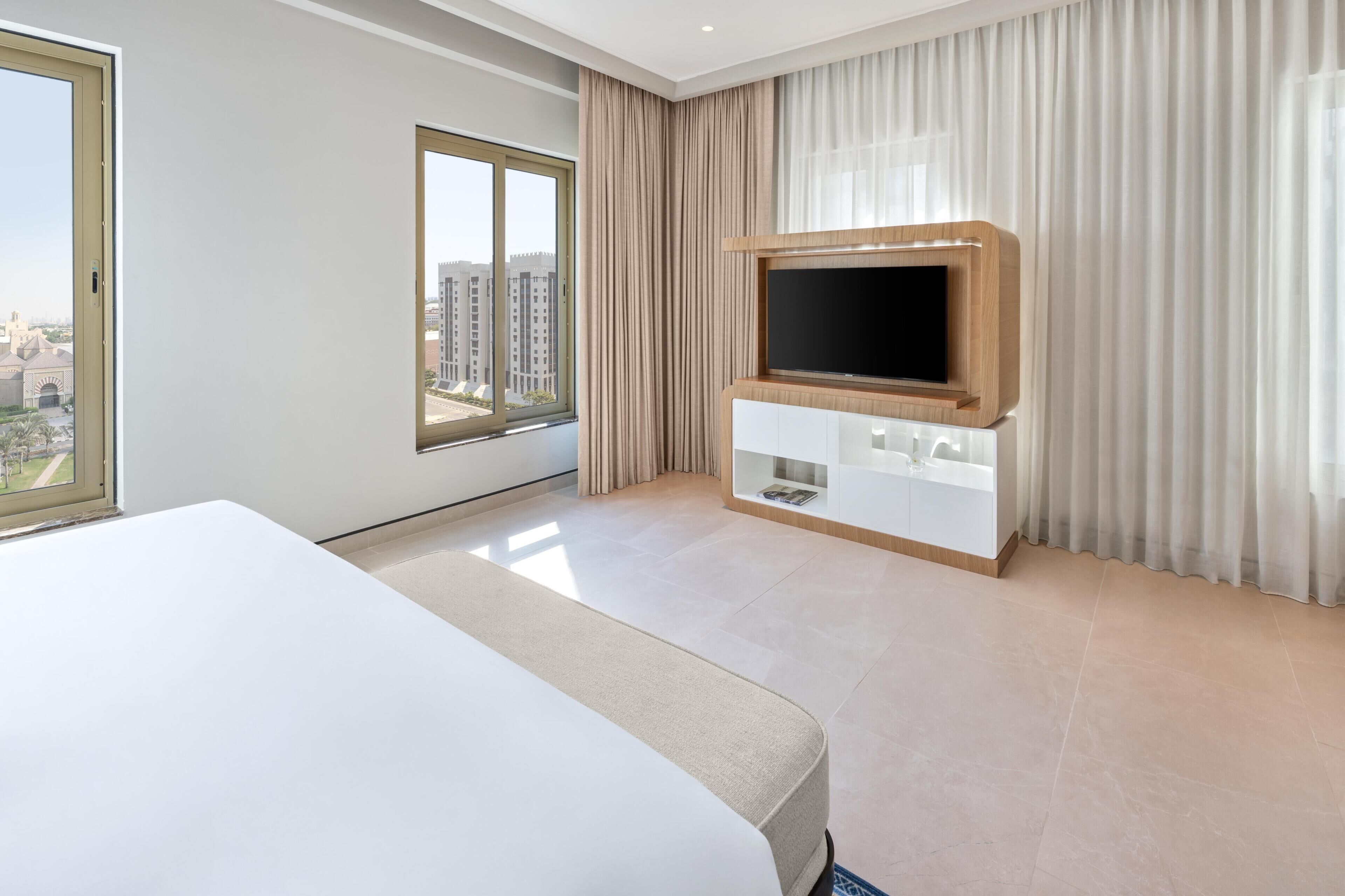 A modern hotel room with a large bed, a TV mounted on a wooden cabinet, a window with curtains, and a view of buildings outside.