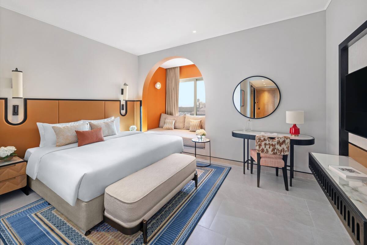 Ein modernes Hotelzimmer mit einem großen Bett, einem Schreibtisch mit Stuhl, einem runden Spiegel und einem Fernseher.