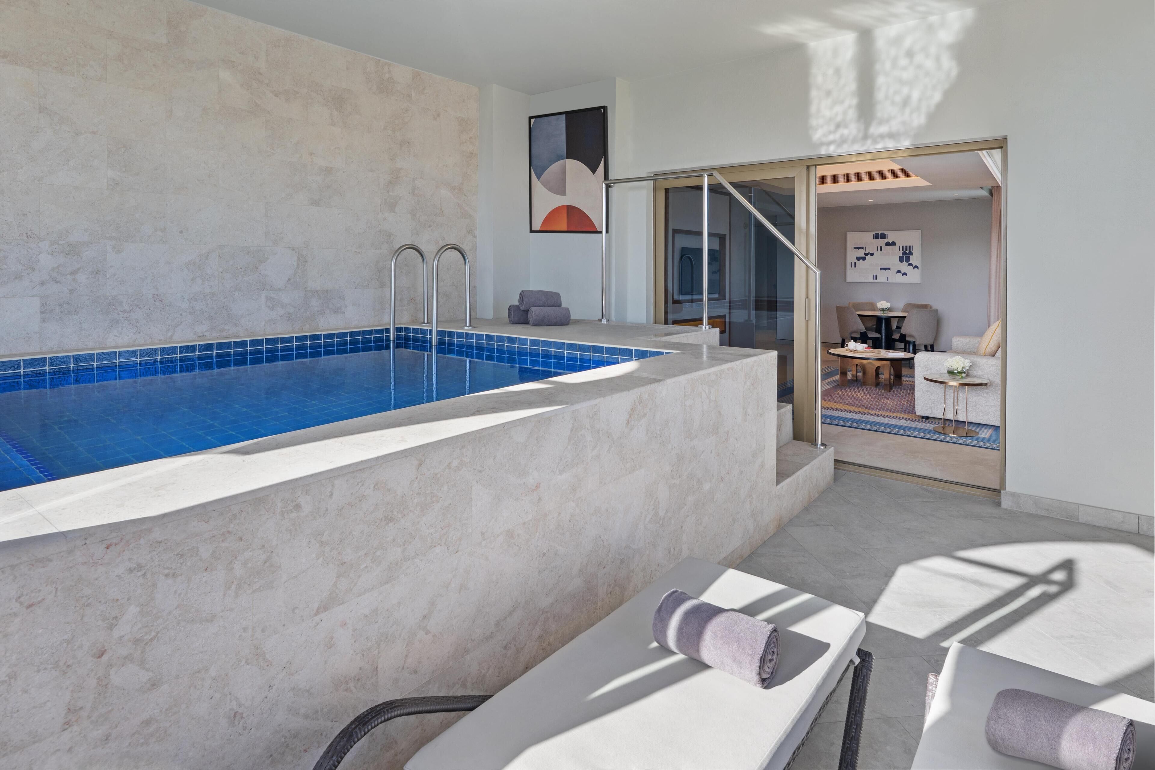 Un espace piscine intérieur moderne, avec une piscine à l’eau cristalline, un espace détente avec des serviettes, et une vue sur un coin repas comprenant une table et des chaises.