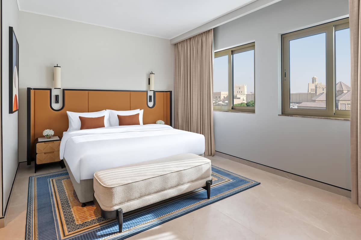 Ein Hotelzimmer mit einem großen Bett, einem Nachttisch, einem Beistelltisch, einem Fenster mit Blick auf die Stadt und einem blauen Teppich mit Muster.