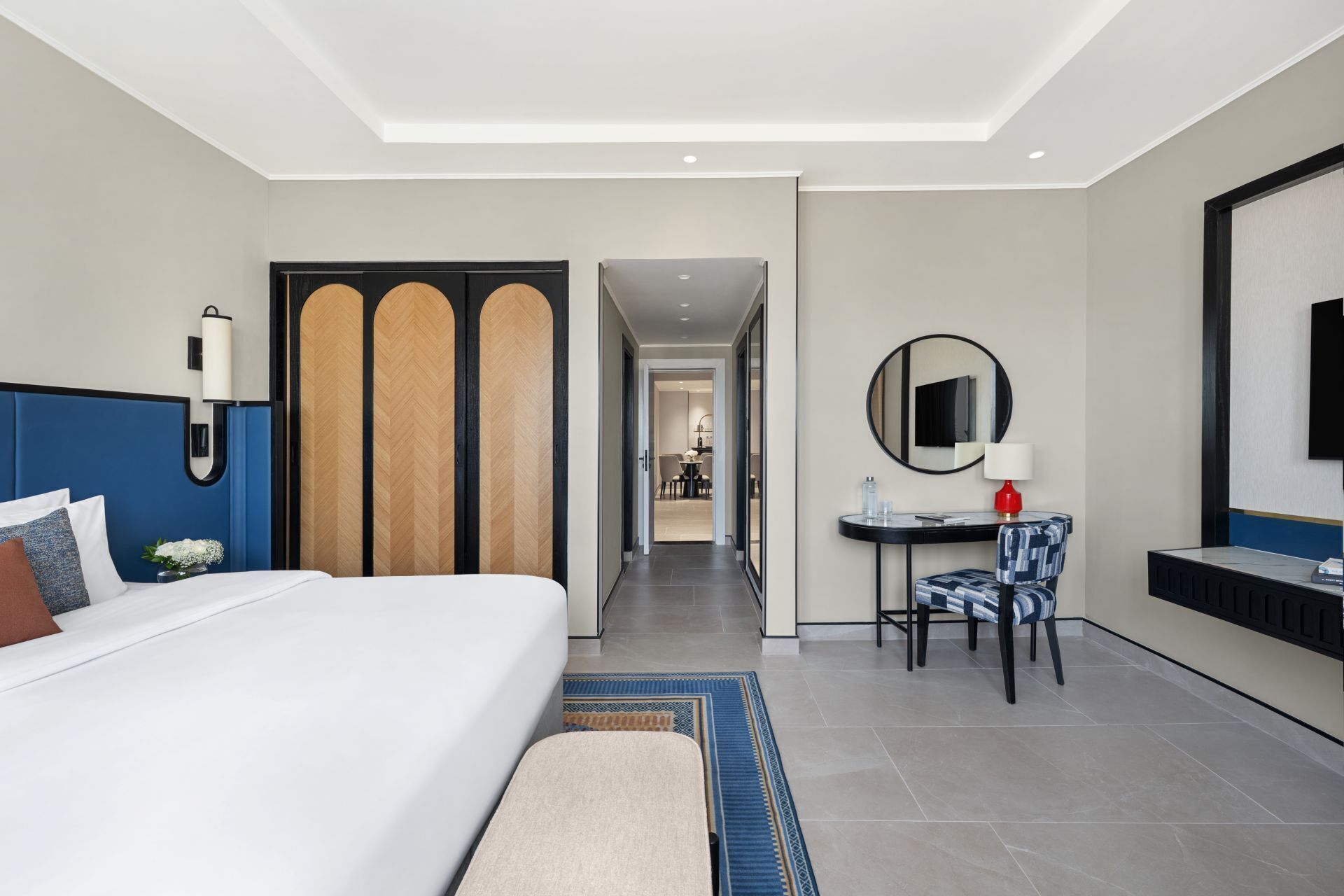 A hotel room with a large bed, a desk with a chair, a mirror, and a view into the hallway.