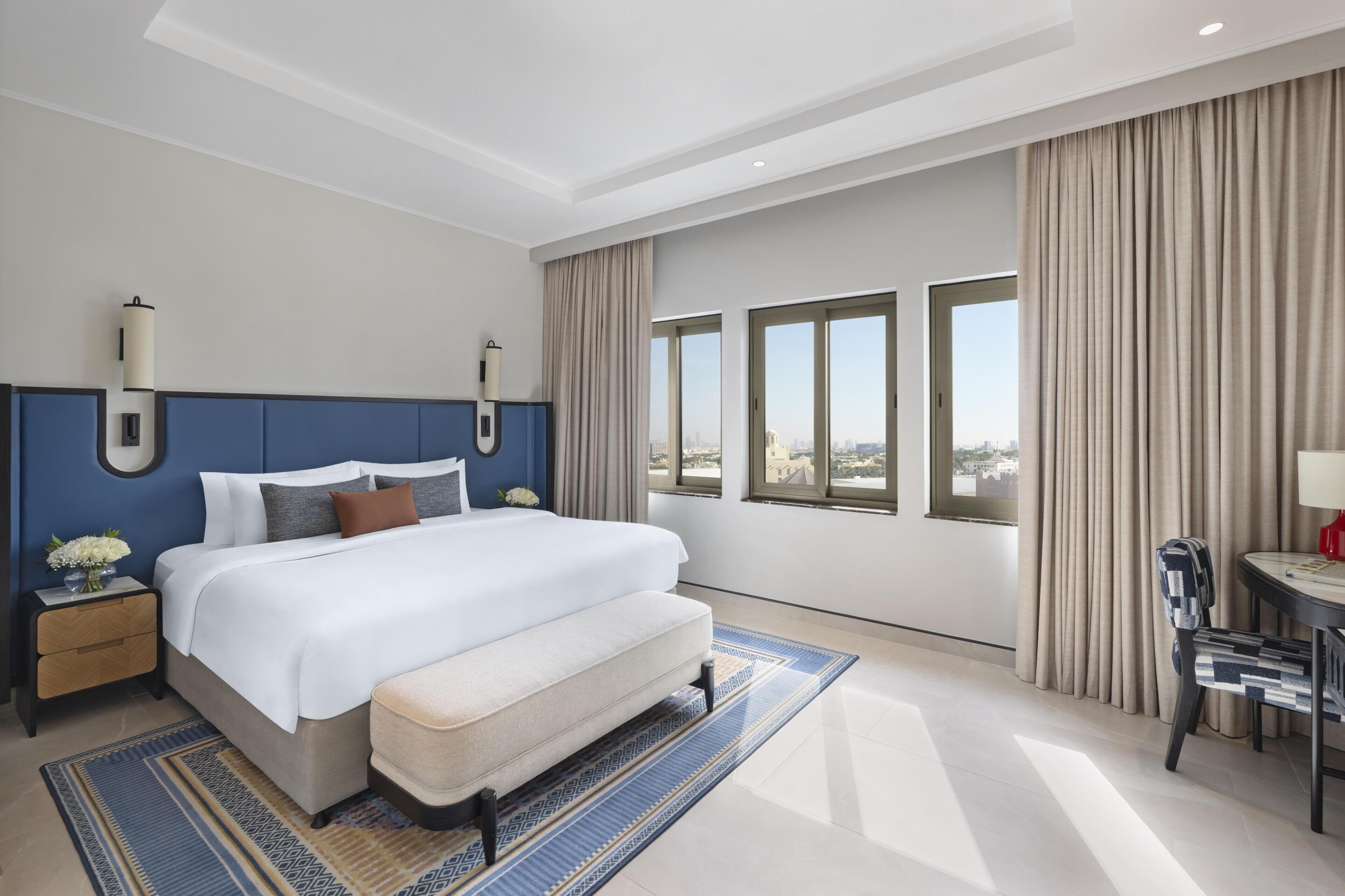 A modern hotel room with a large bed, a bedside table, a chair, a small table with a lamp, and a view of the city through large windows.