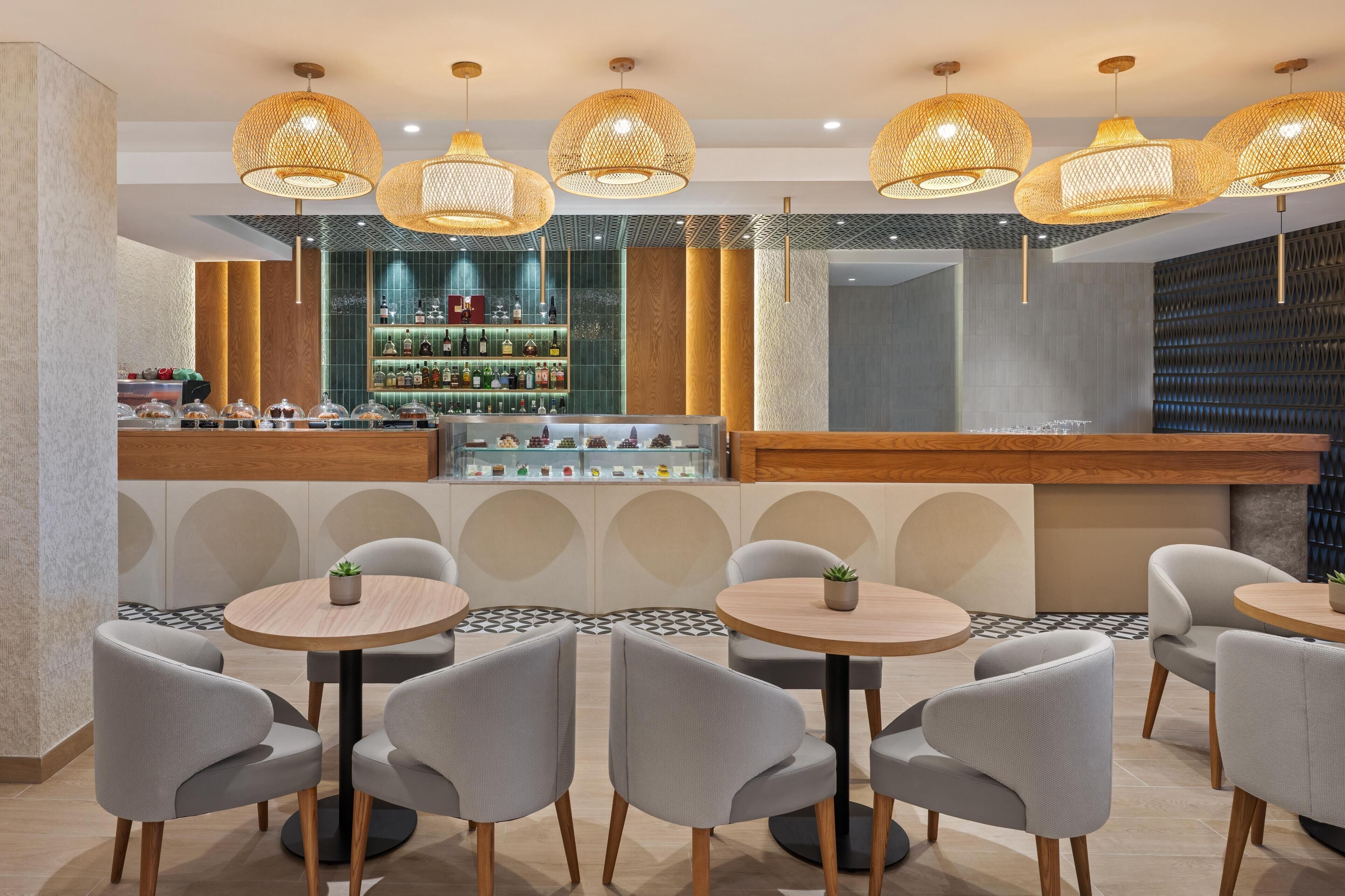 Un espace bar moderne avec des tables et des chaises rondes, un comptoir de bar où sont exposées diverses bouteilles, et des lampes décoratives suspendues.