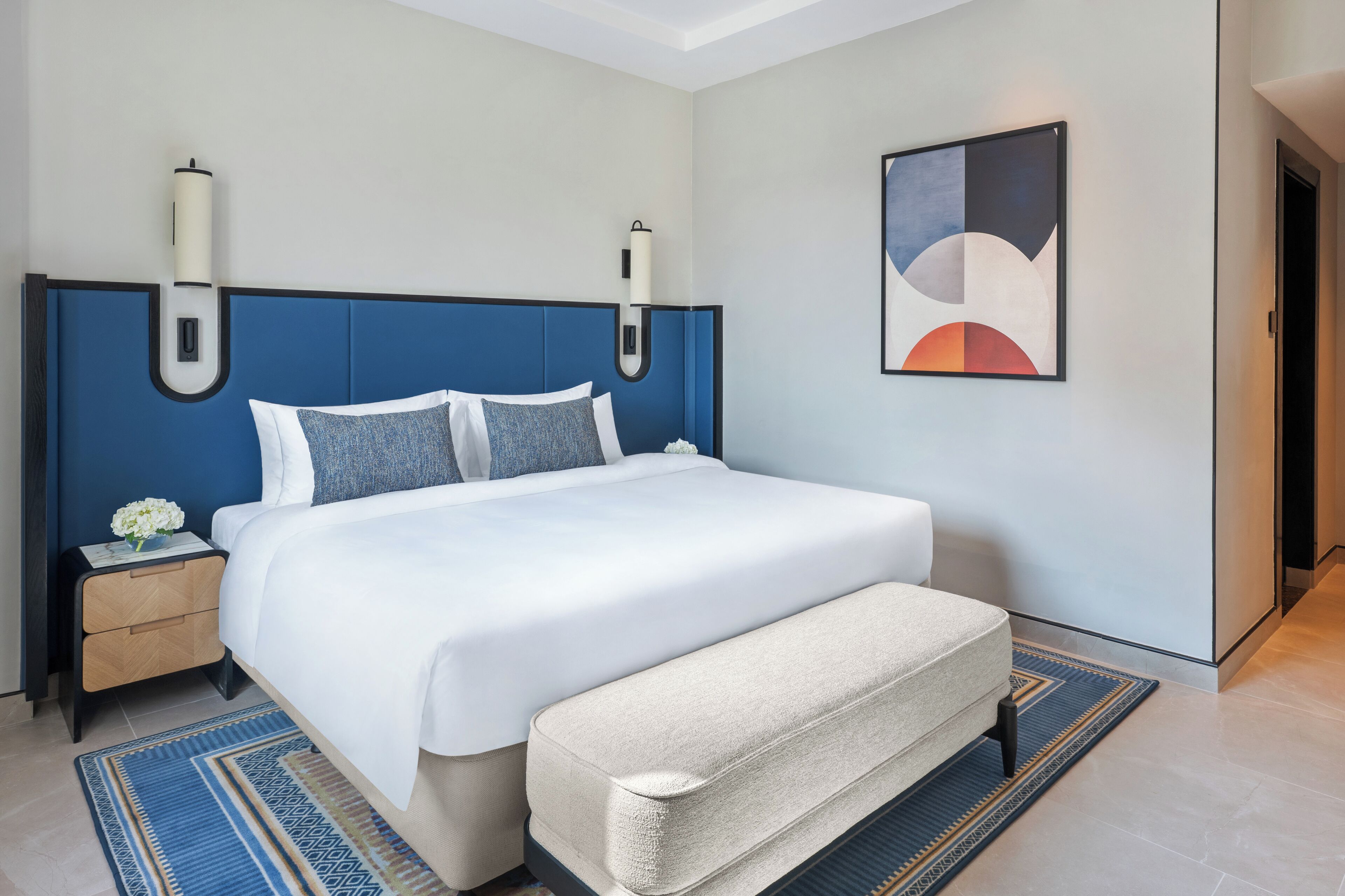 A modern hotel room with a large bed, a bedside table with a vase of flowers, a bench, and a painting on the wall.