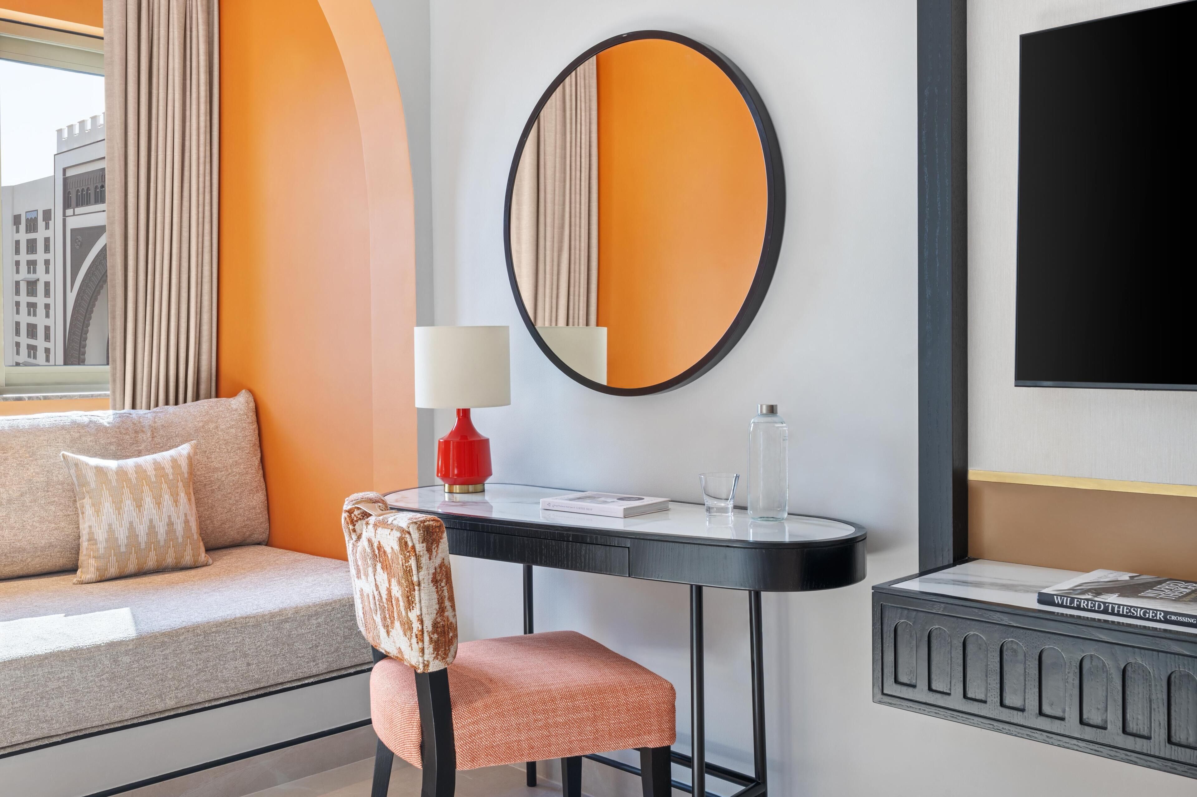 A modern hotel room with a sofa, a chair, a table with a lamp, a mirror, and a TV.