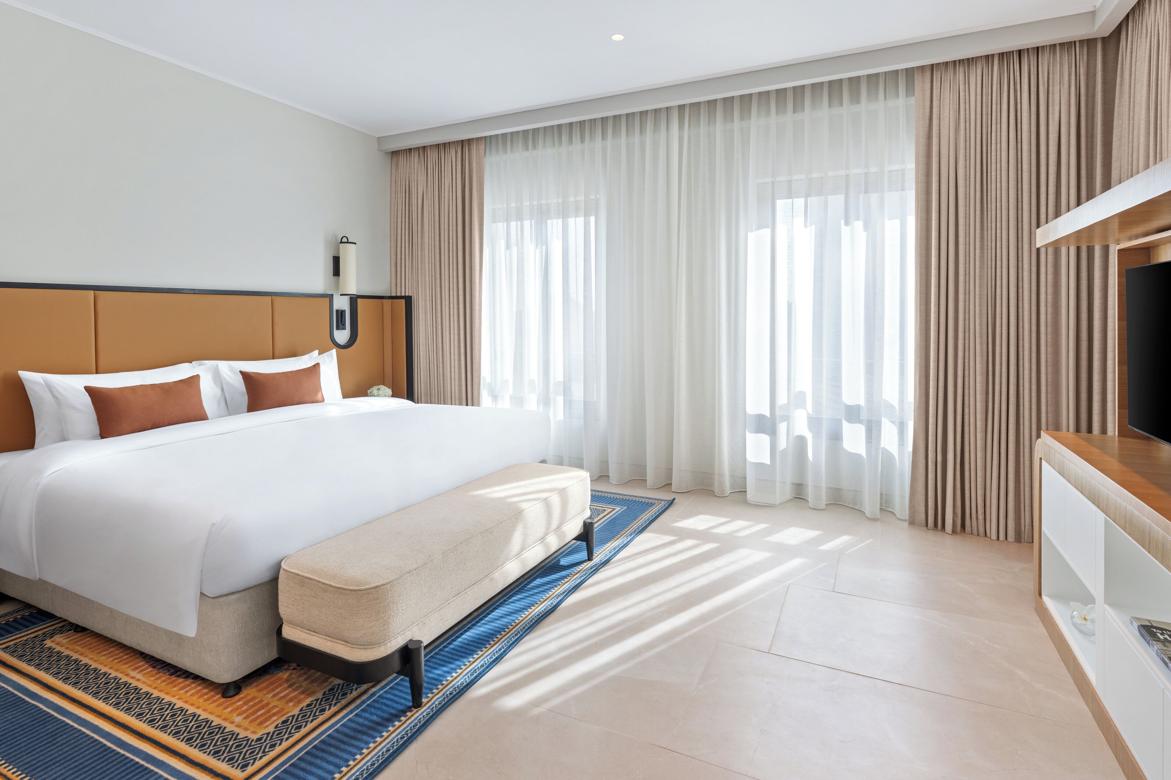 Habitación de hotel moderna con una cama grande, un televisor y vistas de la ciudad a través de cortinas traslúcidas.
