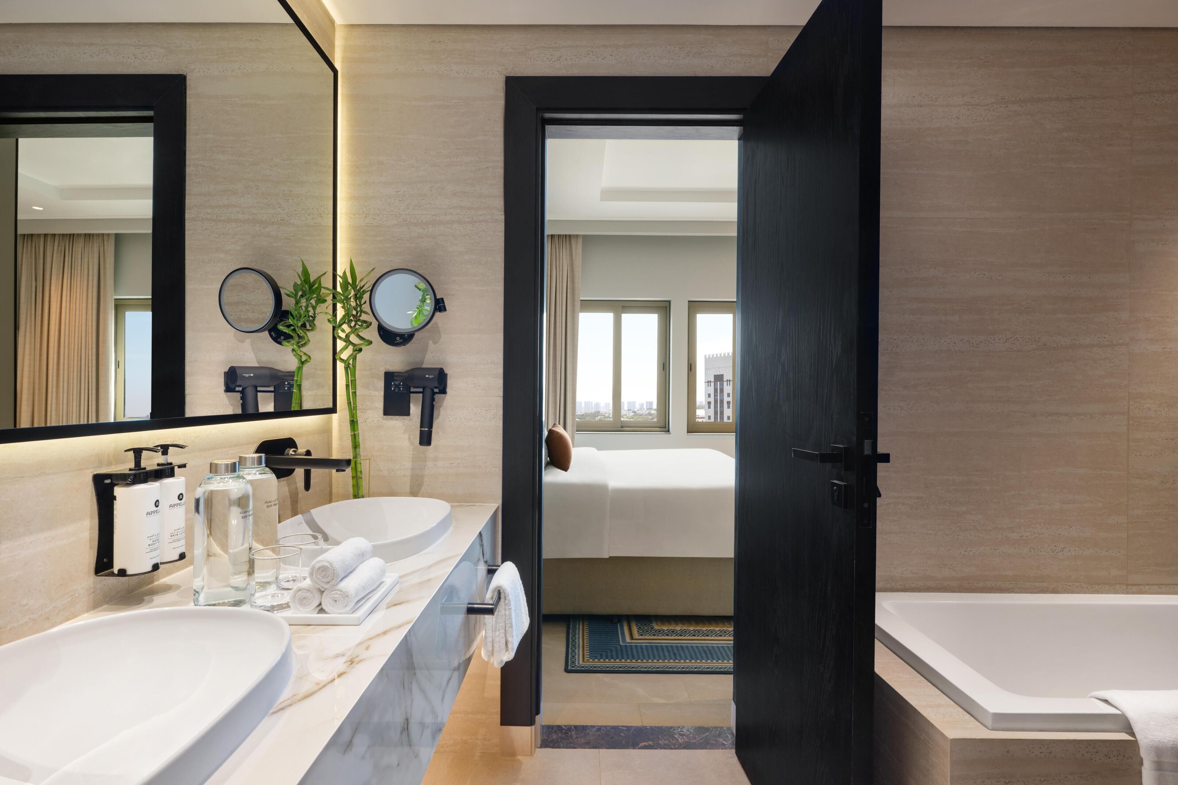 Een moderne badkamer met een grote spiegel, twee wastafels, een bad en een deur naar een slaapkamer.