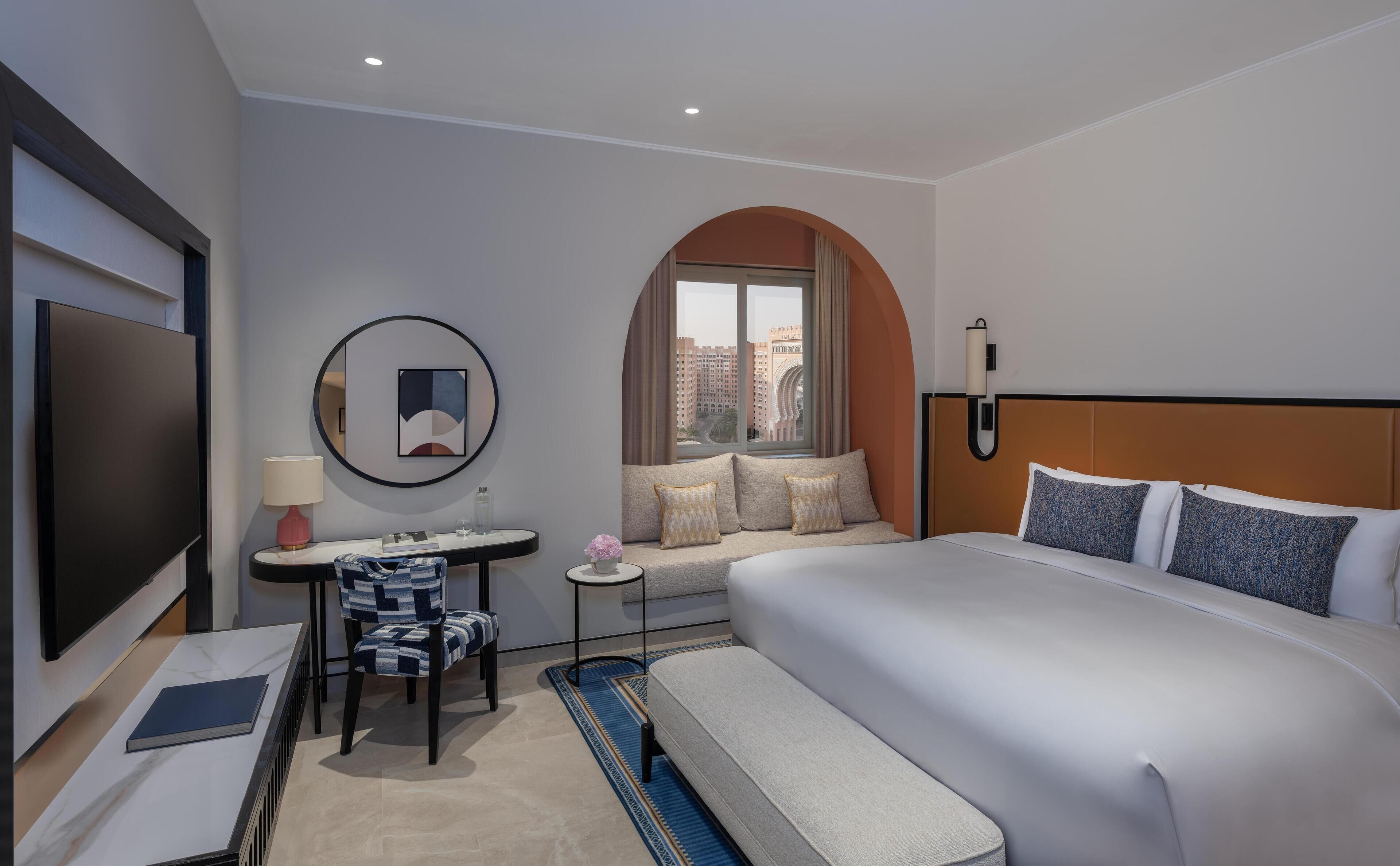 Ein modernes Hotelzimmer mit einem großen Bett, einem Schreibtisch mit Stuhl, einem kleinen Tisch mit Lampe und Blick auf die Stadt durch ein rundbogenförmiges Fenster.