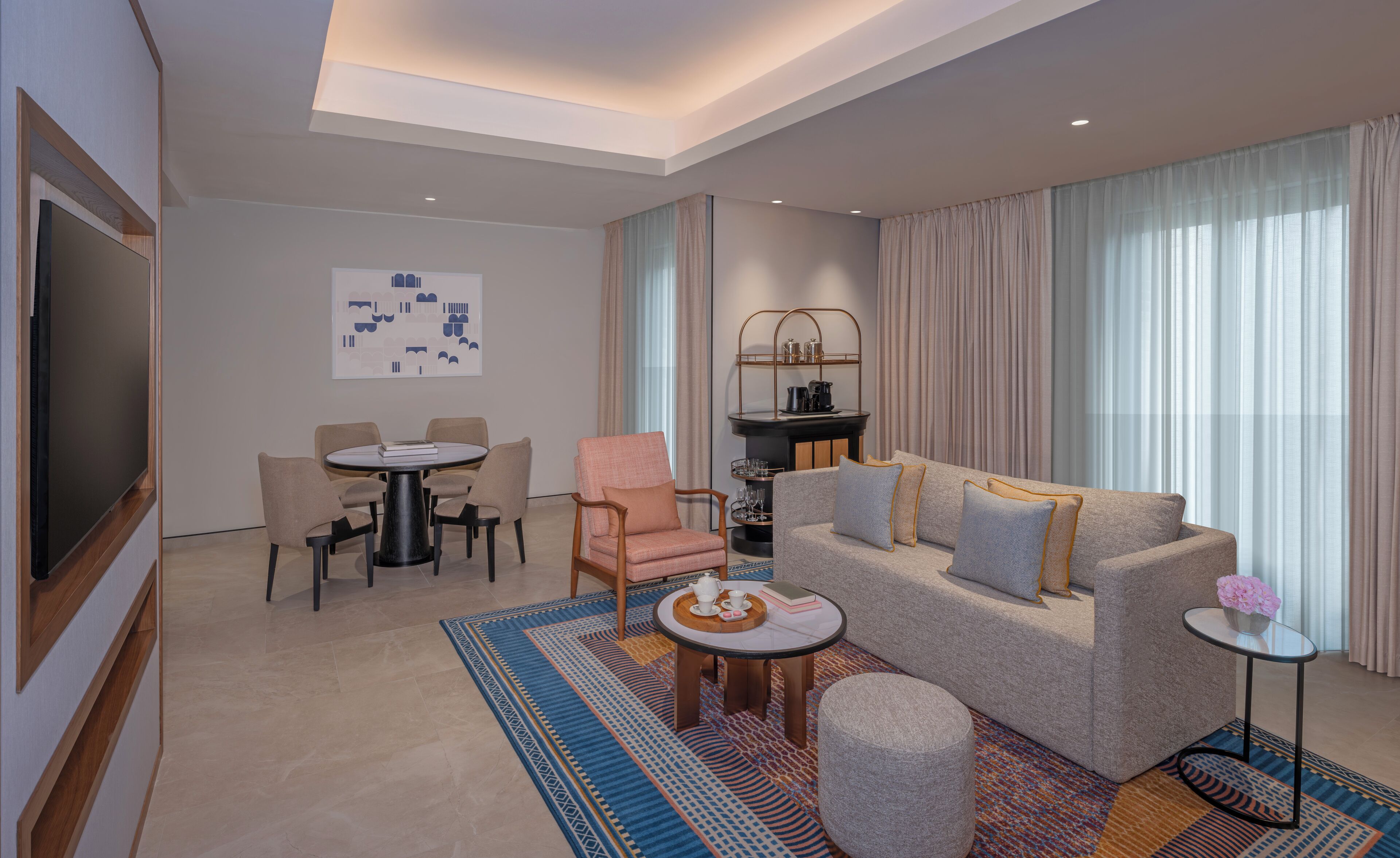 A modern hotel room with a flat-screen TV, a dining area with a round table and chairs, a sofa with cushions, and a coffee table with a tray.