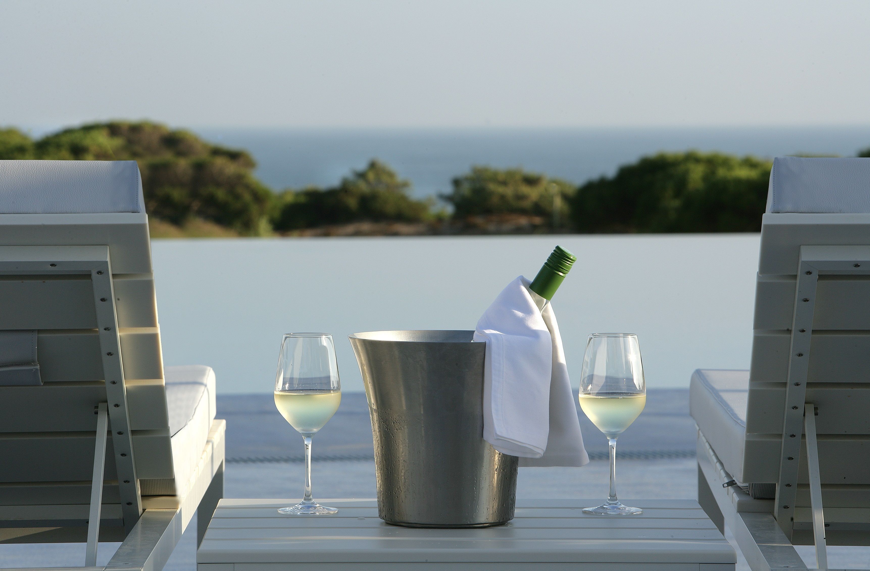 A table with a champagne bottle, two glasses, and a bucket, overlooking a scenic view of the sea and greenery.