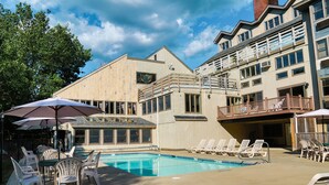 A pool area with lounge chairs and umbrellas, a dining table with chairs, and a building with multiple windows and balconies. - The Valley Inn, an Ascend Collection Hotel (Waterville Valley)