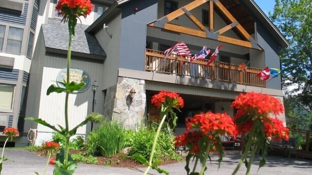 A hotel with a balcony decorated with flags, a stone facade, and red flowers in the foreground. - The Valley Inn, an Ascend Collection Hotel (Waterville Valley)