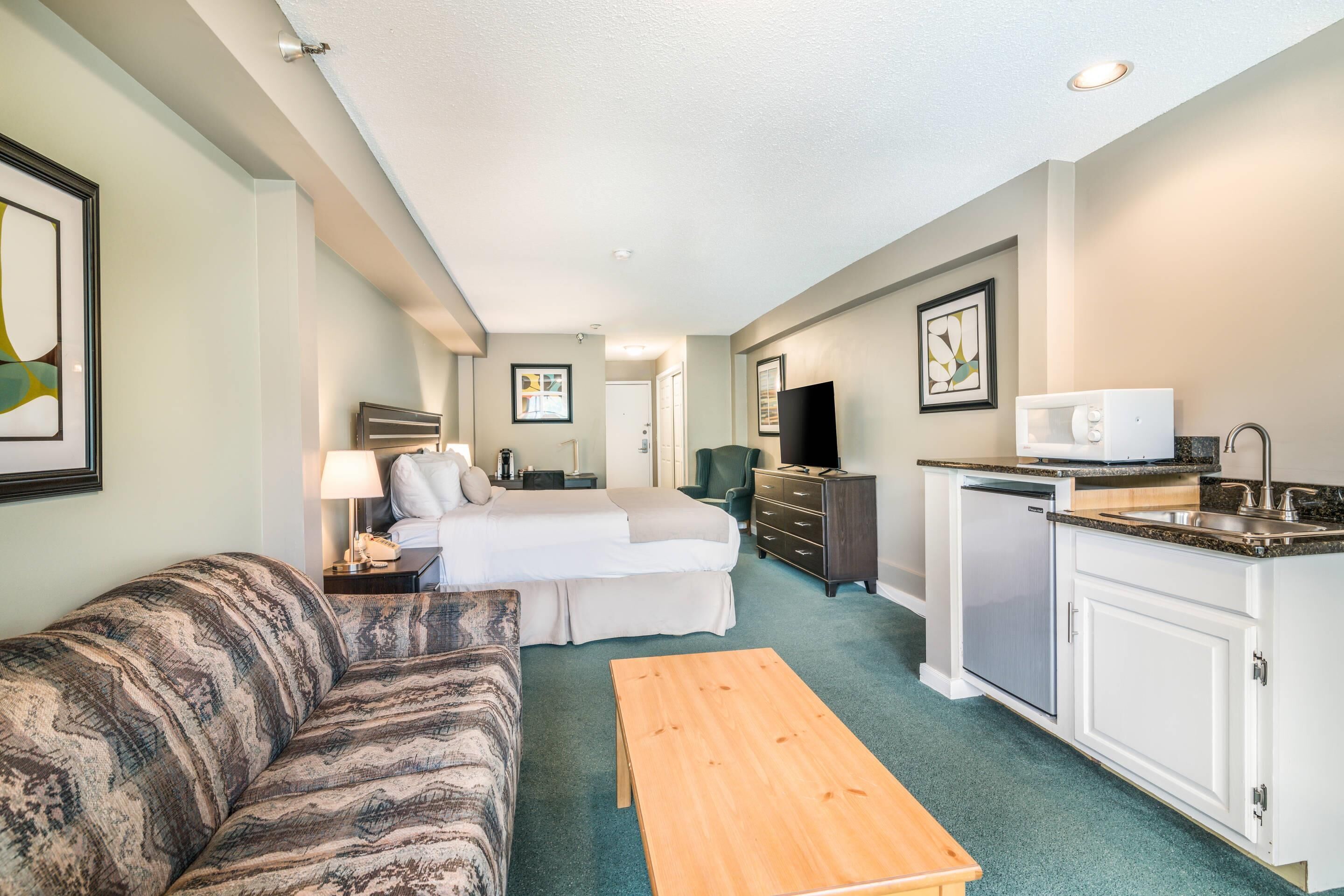 A compact hotel room with a bed, sofa, kitchenette, and a small dining table.