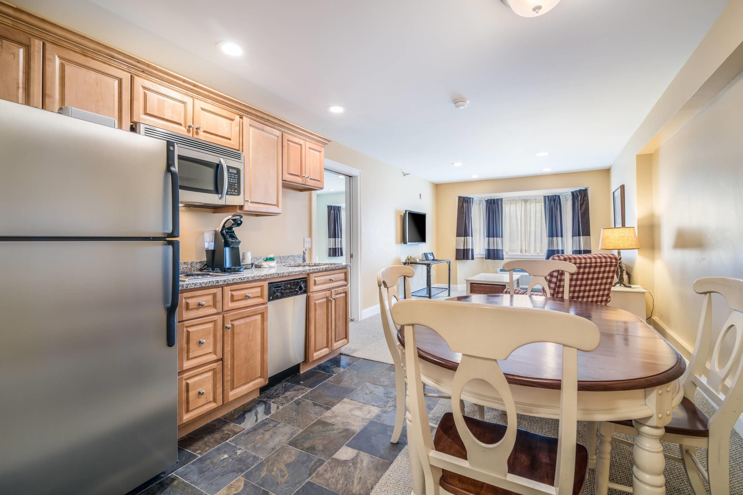 A kitchen with wooden cabinets, a stainless steel refrigerator, a microwave, and a dishwasher. There is a dining table with chairs and a lamp.