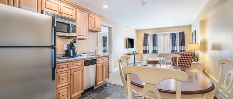 A kitchen with wooden cabinets, a stainless steel refrigerator, a microwave, and a dishwasher. There is a dining table with chairs and a lamp.