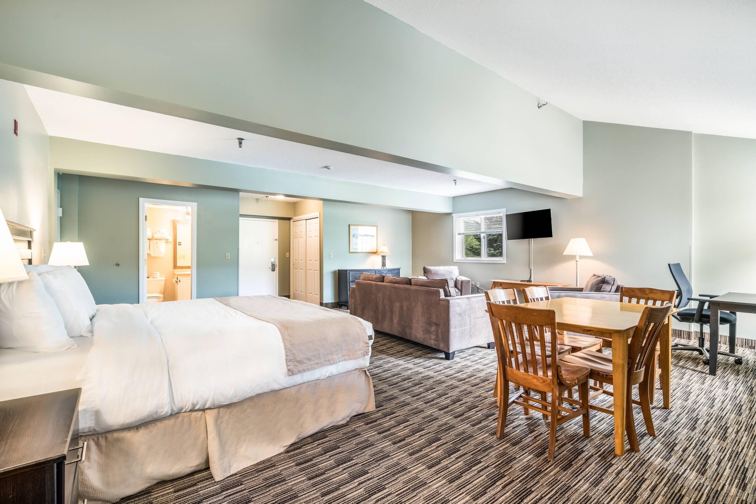 A hotel room with a bed, a sofa, a dining table with chairs, a TV, and a lamp.