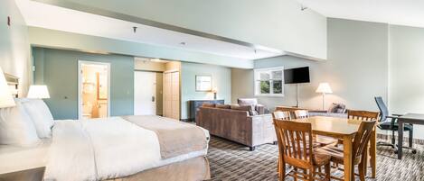 A hotel room with a bed, a sofa, a dining table with chairs, a TV, and a lamp.