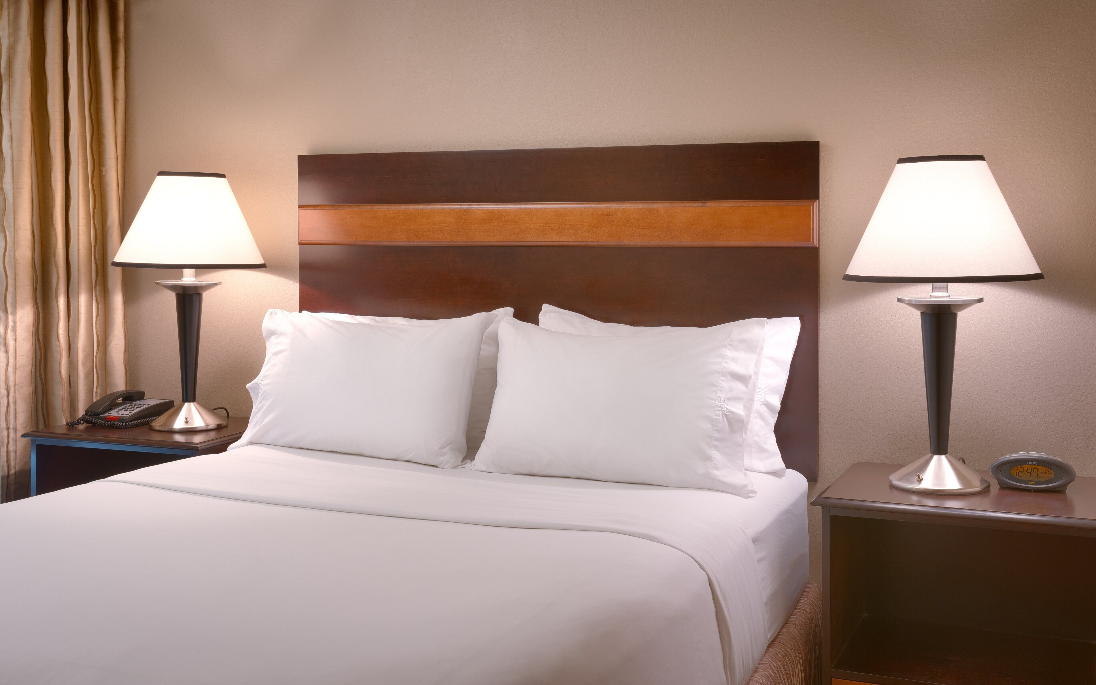 A hotel room with a bed, two bedside lamps, a nightstand with a phone, and a digital clock.