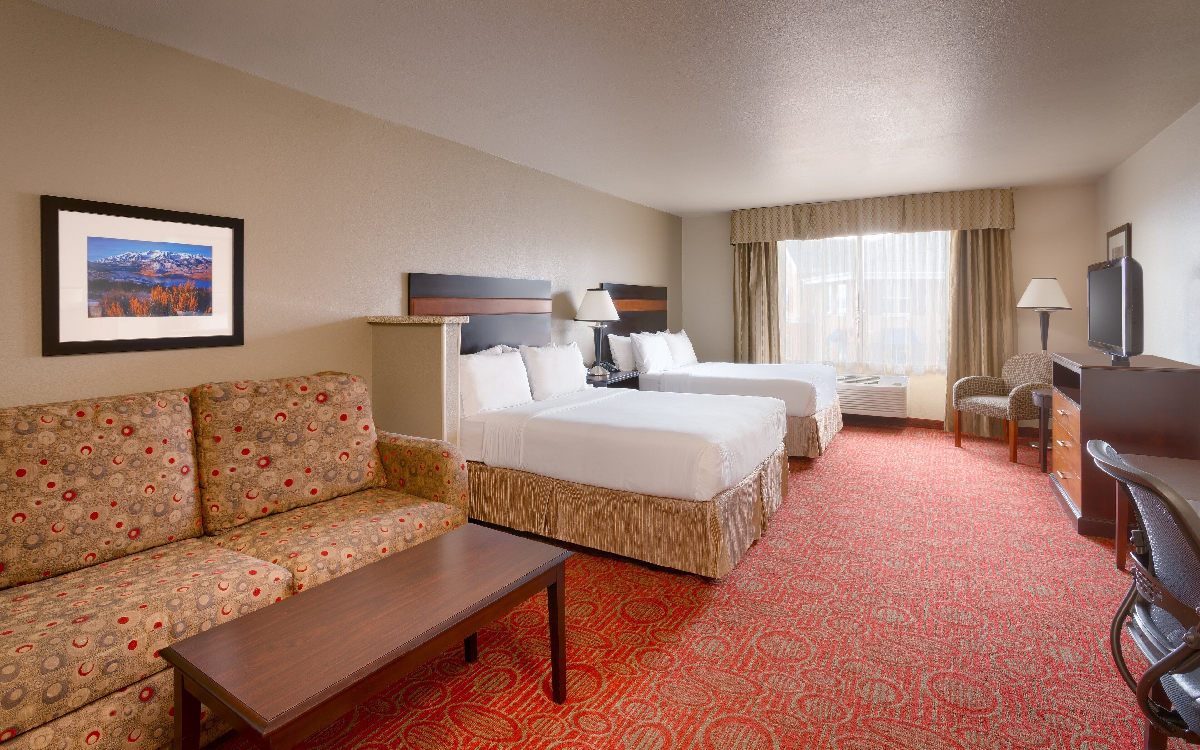 A hotel room with a sofa, two beds, a desk, a TV, and a framed picture on the wall.