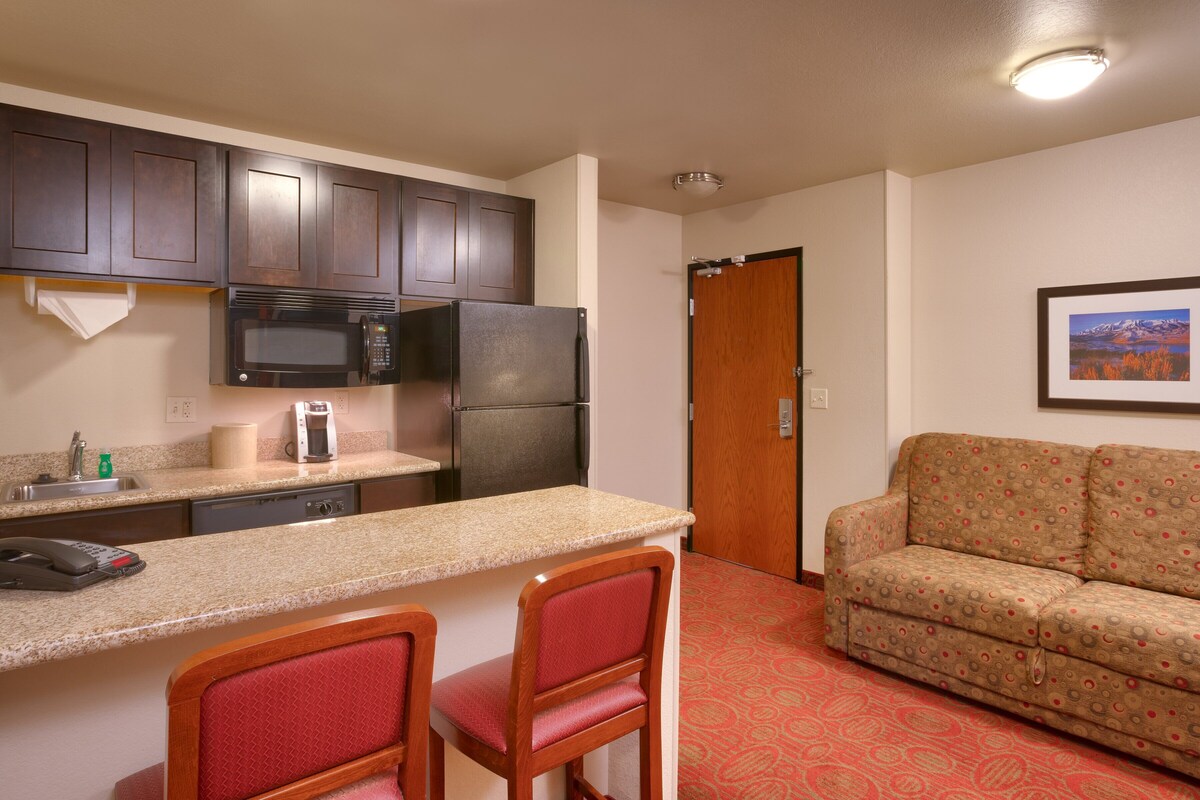 A compact kitchen with a microwave, refrigerator, and coffee maker. There is a wooden door leading to a living area with a sofa and a framed picture on the wall.