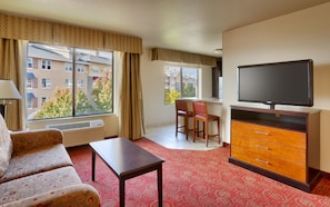 A hotel room with a sofa, a small table, a TV on a wooden cabinet, a chair, and a large window with curtains. - Holiday Inn Express & Suites Orem-North Provo by IHG (Orem)