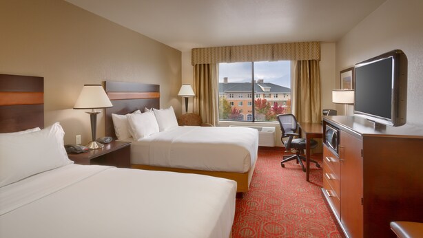 A hotel room with two beds, a desk with a computer, a TV, and a window with a view of buildings and trees. - Holiday Inn Express & Suites Orem-North Provo by IHG (Orem)