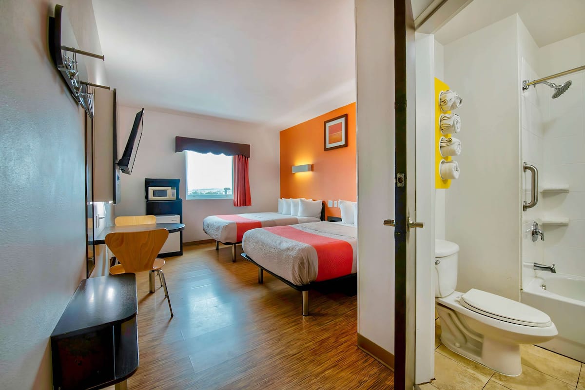 A hotel room with two beds, a desk, a chair, a window with curtains, a bathroom with a toilet and shower, and a wall-mounted towel rack.