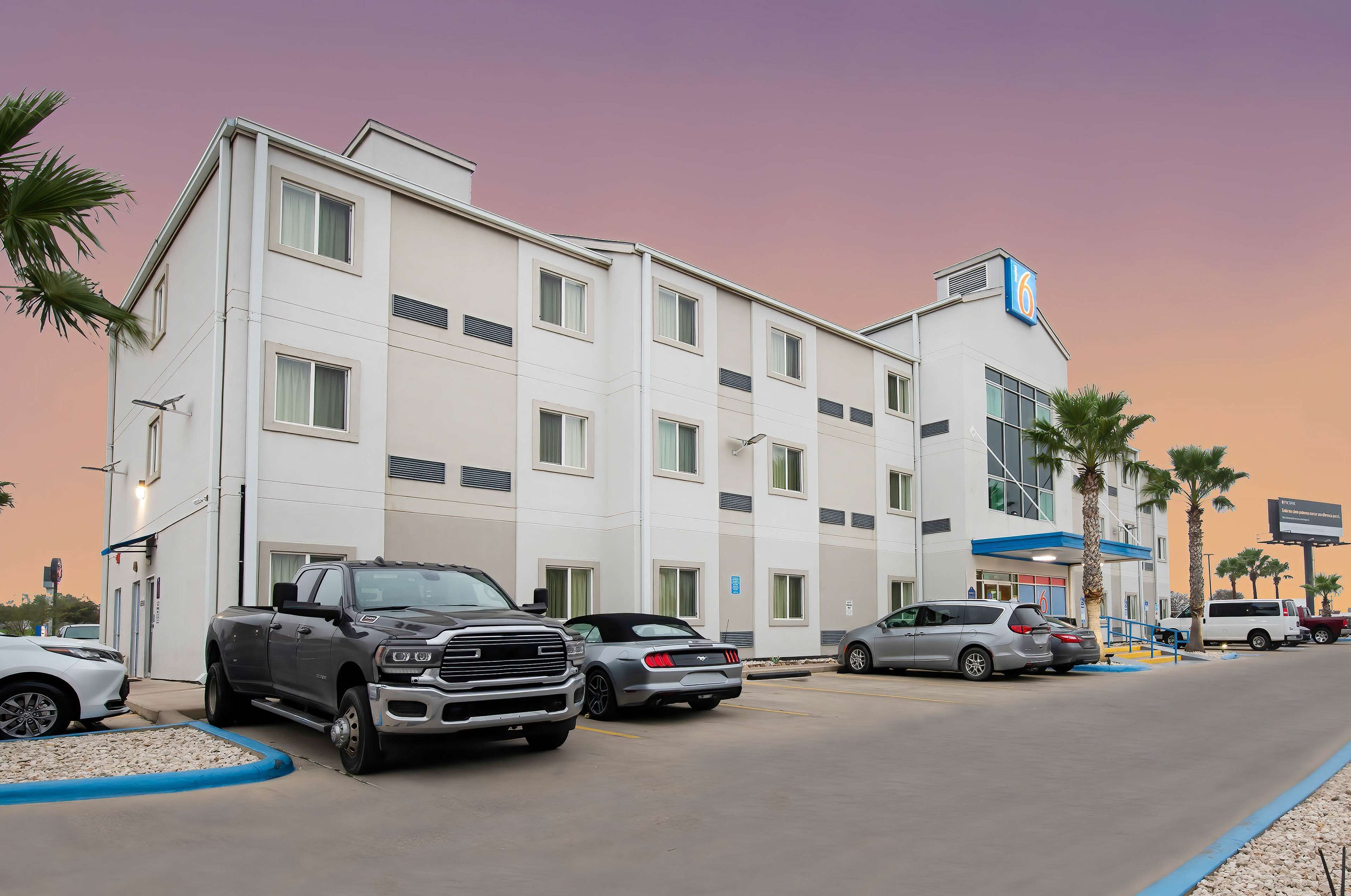 A multi-story hotel with a flat facade, a prominent '6' sign, and a parking lot with various vehicles.