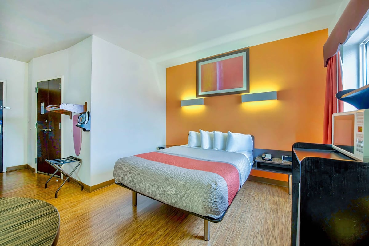 A hotel room with a bed, bedside table, and a desk with a chair. There is a door and a window with curtains.