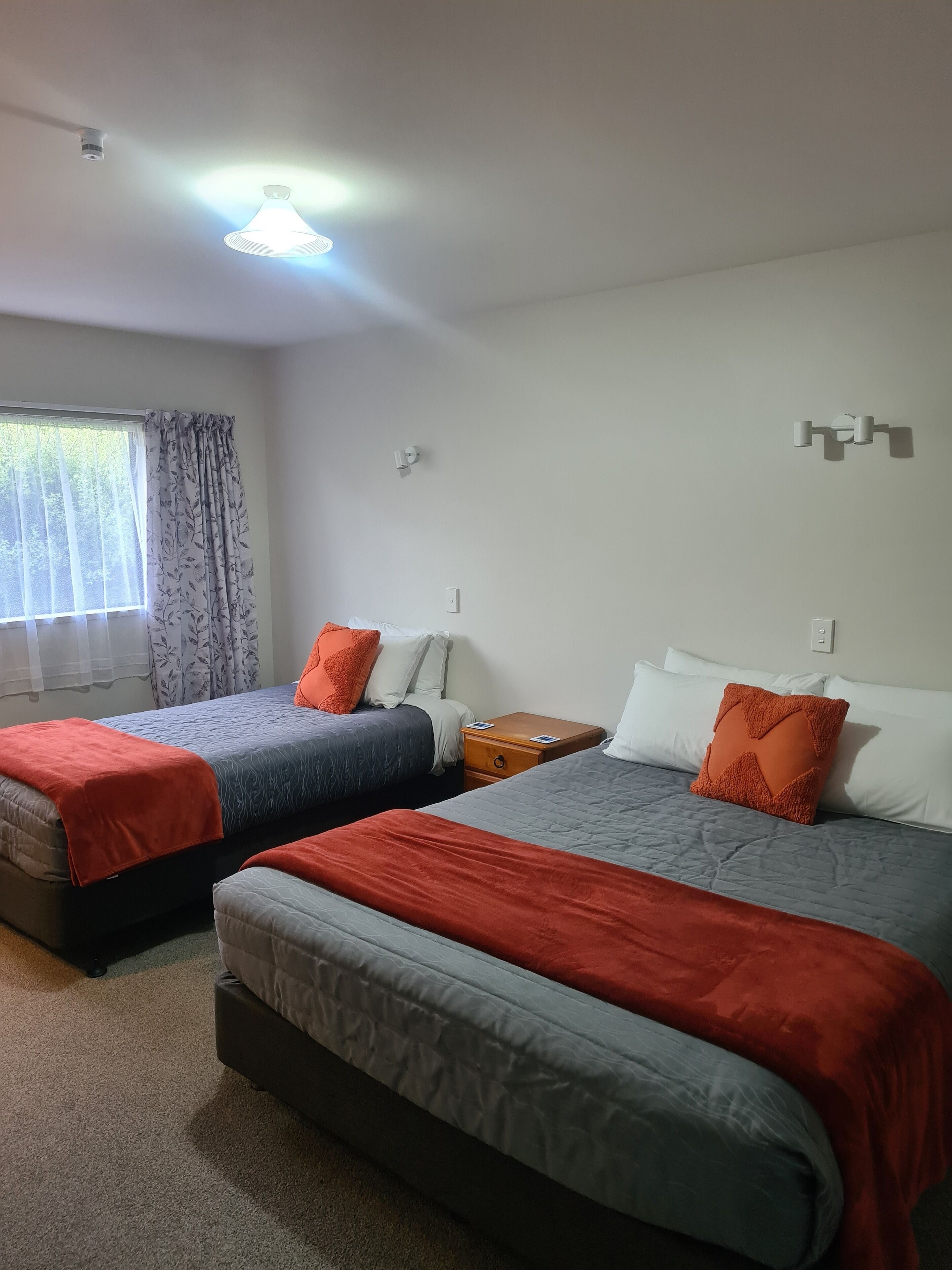 A hotel room with two beds, a window with sheer curtains, a ceiling light, and a wall-mounted light fixture.