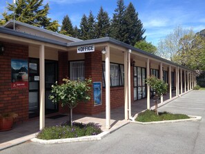 Un edificio de una planta con un cartel que indica una oficina. Hay dos plantas en macetas al frente, un estacionamiento y un cielo despejado.