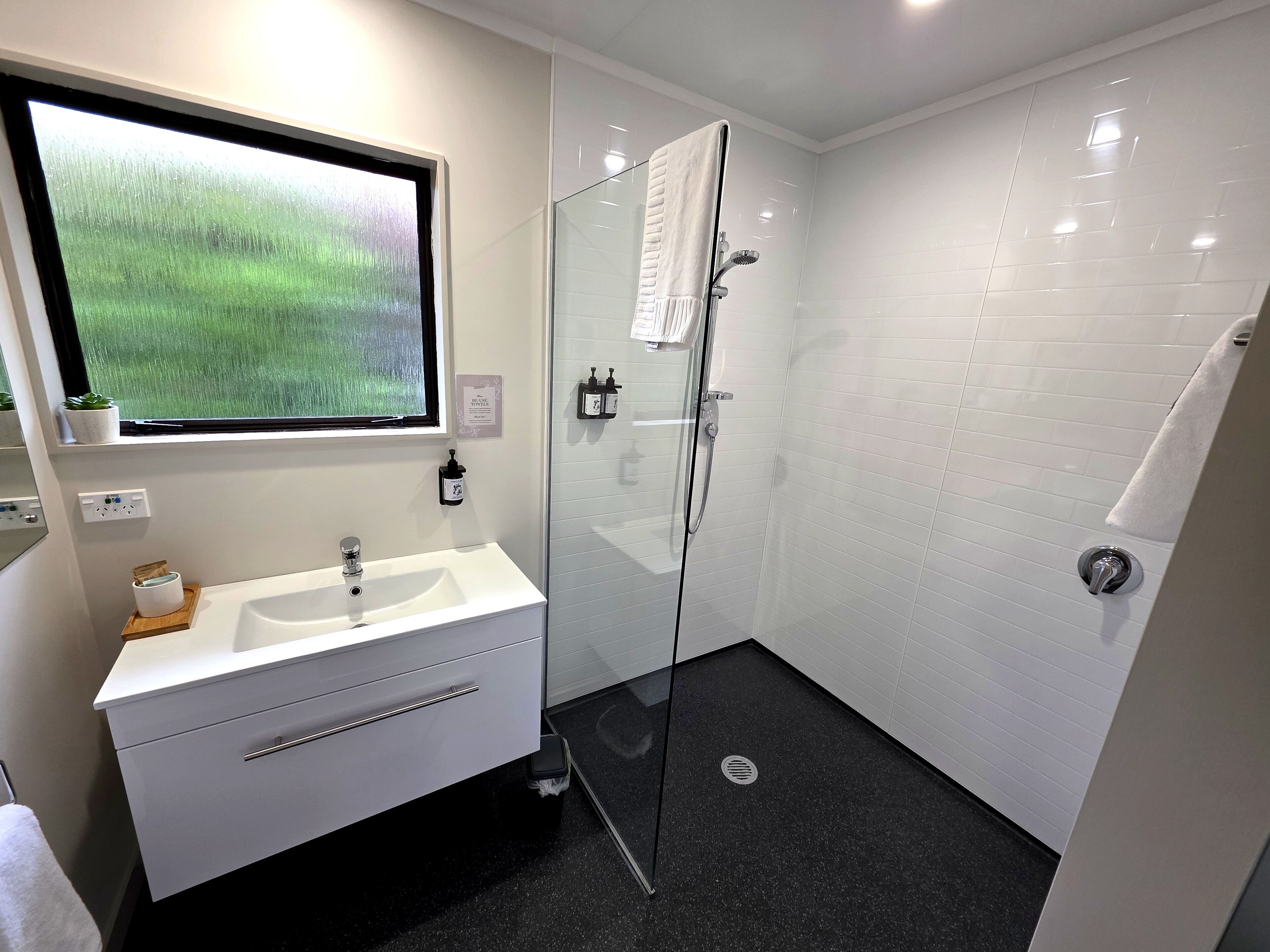 A modern bathroom with a glass shower enclosure, a white sink, and a window with a view of greenery.