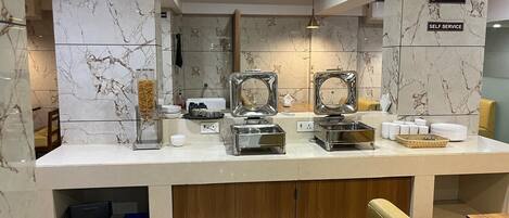 A self-service breakfast area with marble-tiled walls, stainless steel appliances, and a wooden counter.