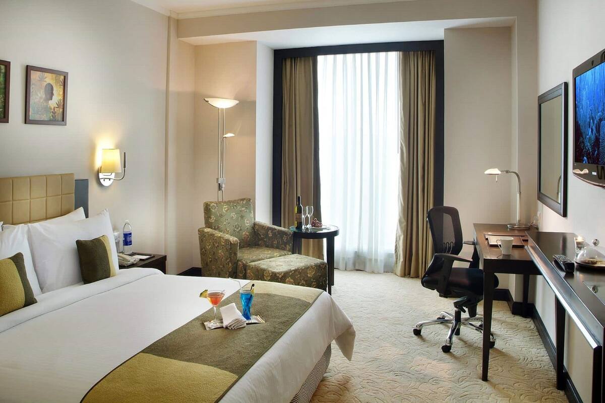Ein Hotelzimmer mit einem großen Bett, einem Schreibtisch, einem Sessel, einer Schreibtischlampe, einem Fernseher und einem Fenster mit Vorhängen.