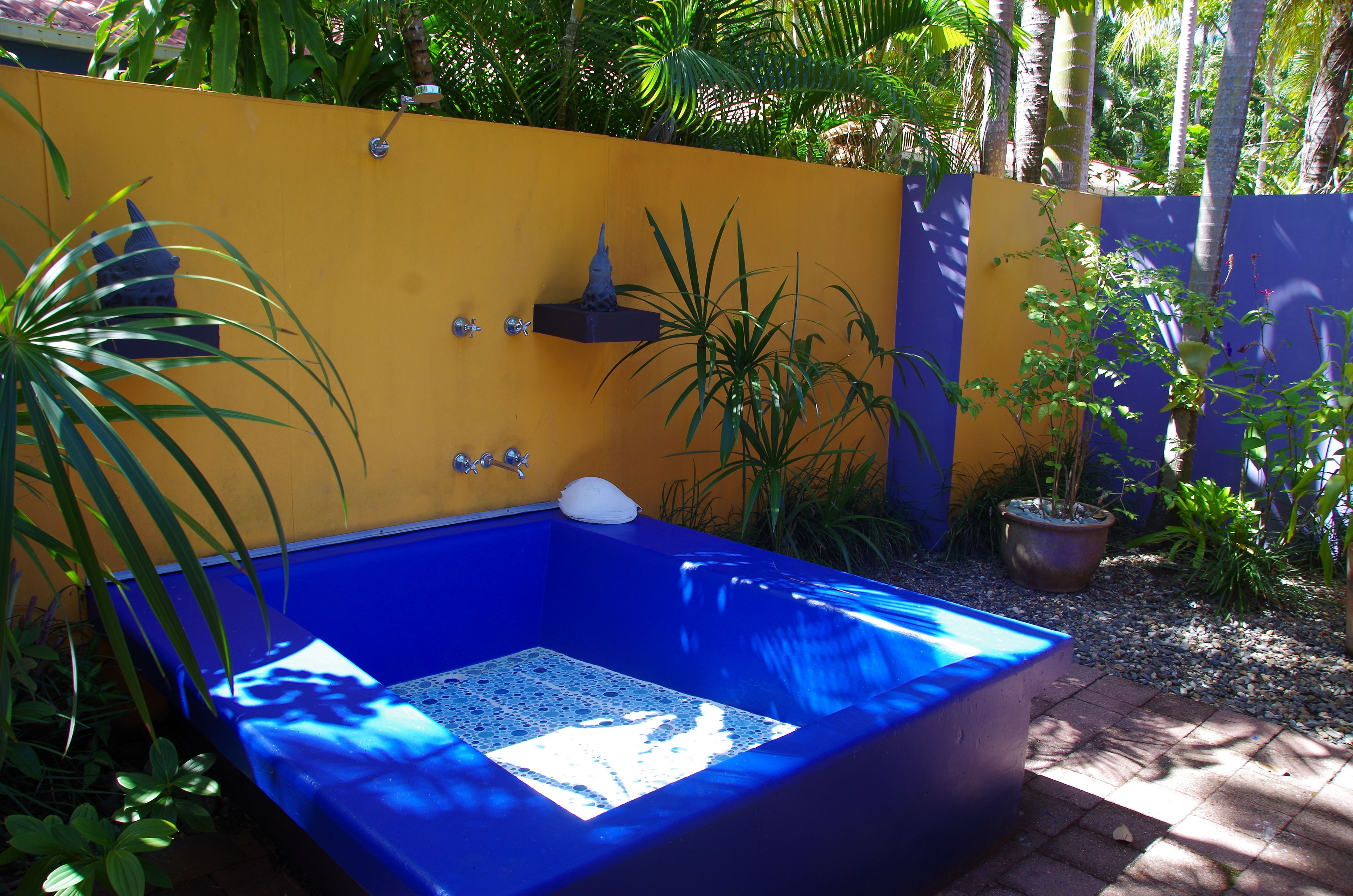 Un bain à remous bleu, avec un sol recouvert de mosaïques blanches, entouré d’un mur jaune et violet, et agrémenté de différentes plantes en pot.