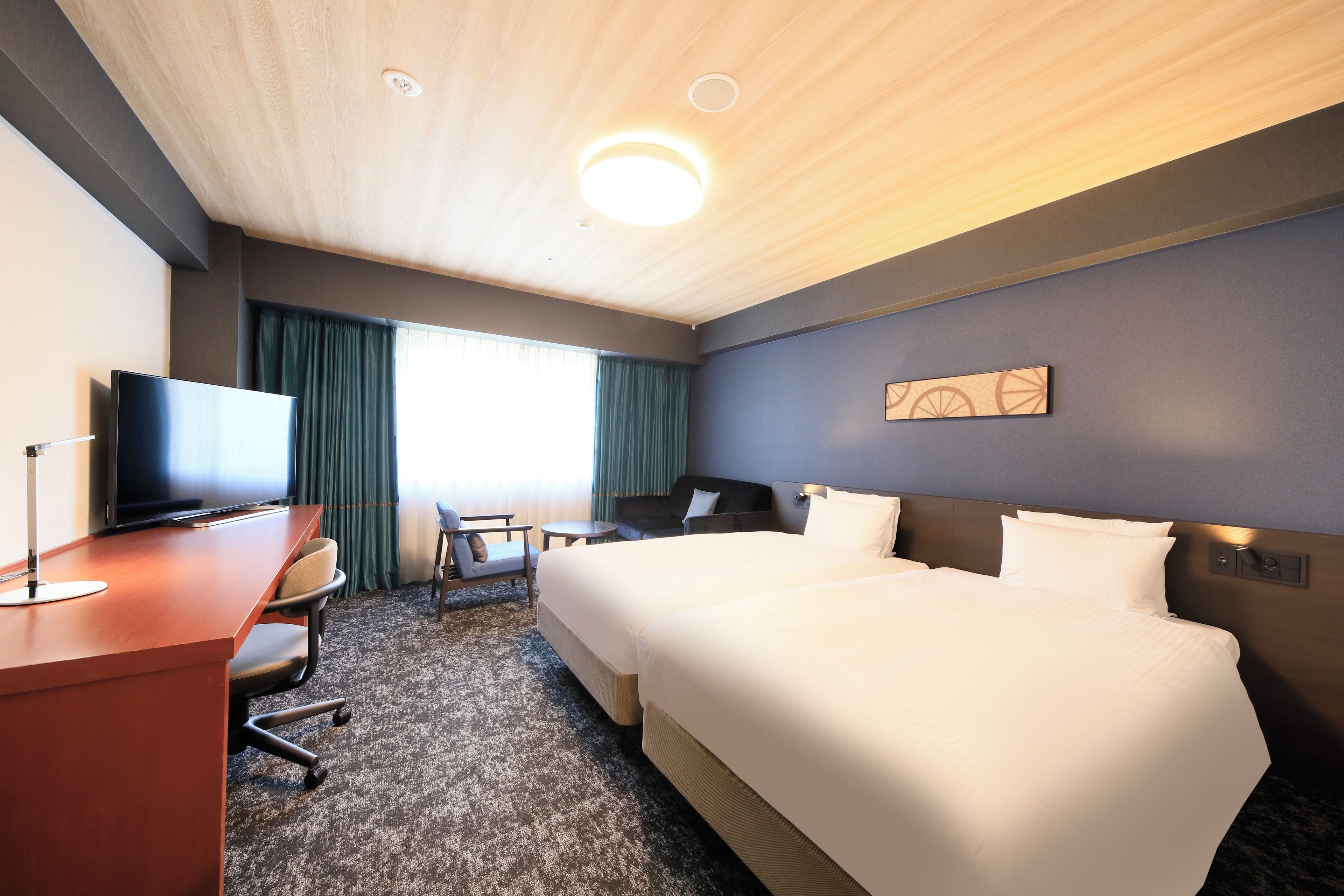 A hotel room with a bed, a desk with a television, a chair, a small table, and a window with curtains.