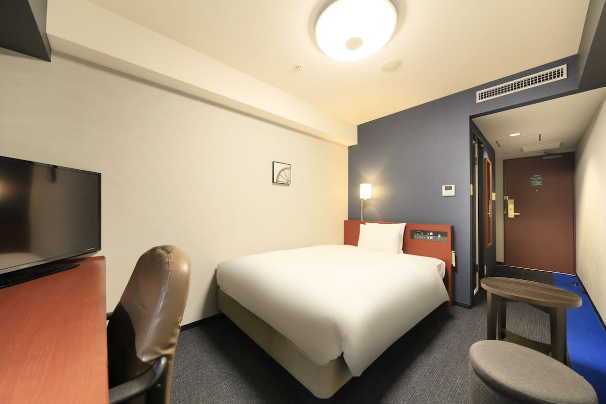 A hotel room with a bed, a desk, a chair, a TV, and a small table.