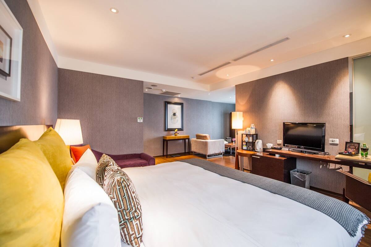 Una habitación de hotel moderna con una cama grande, un escritorio con televisor de pantalla plana, una silla, una mesita y un cesto de basura.