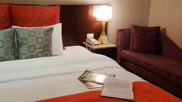 A hotel room with a bed, a nightstand, a lamp, a reading material, and a couch.