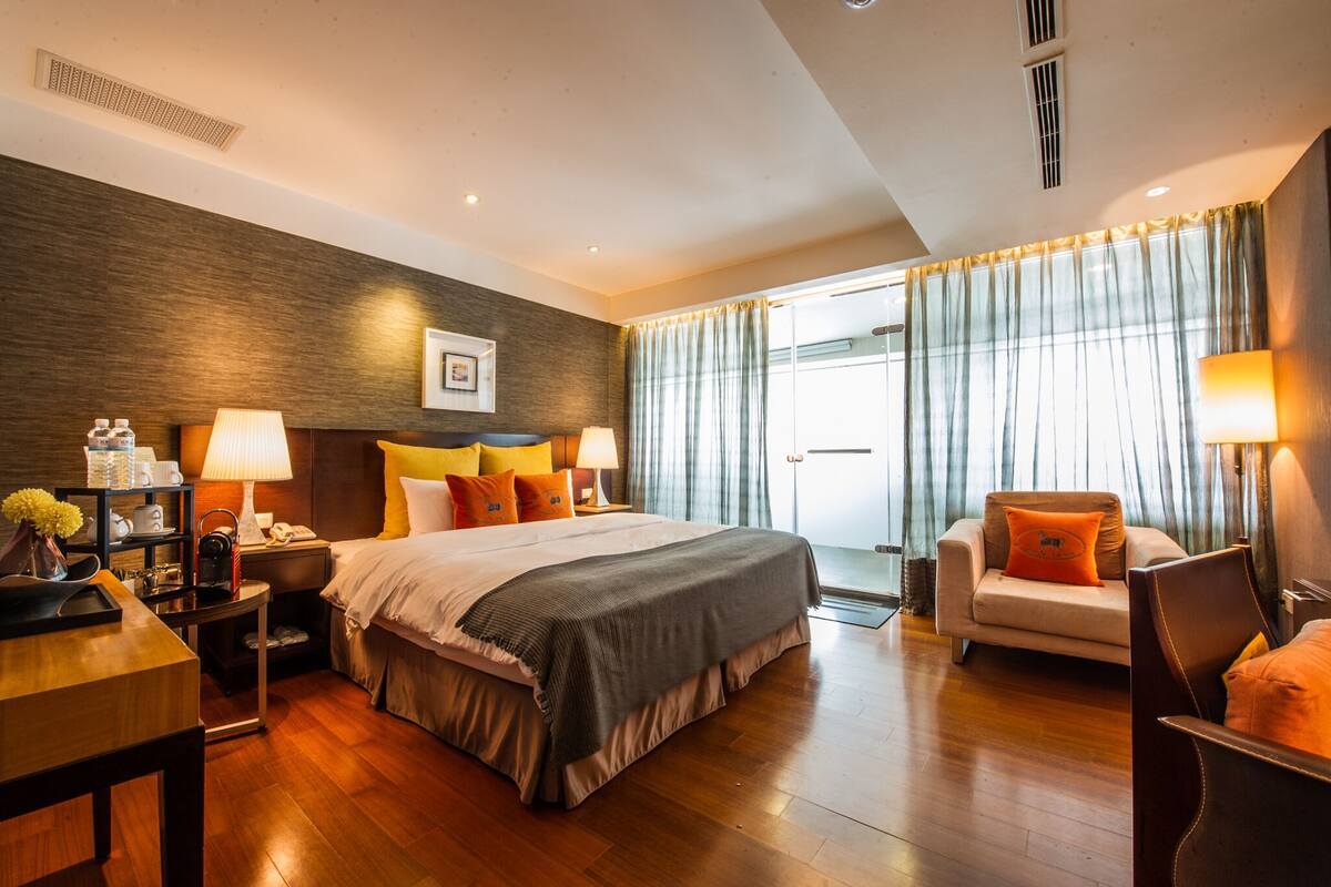 Habitación de hotel con una cama grande, mesitas de noche, un escritorio, una silla y vistas al exterior a través de cortinas.