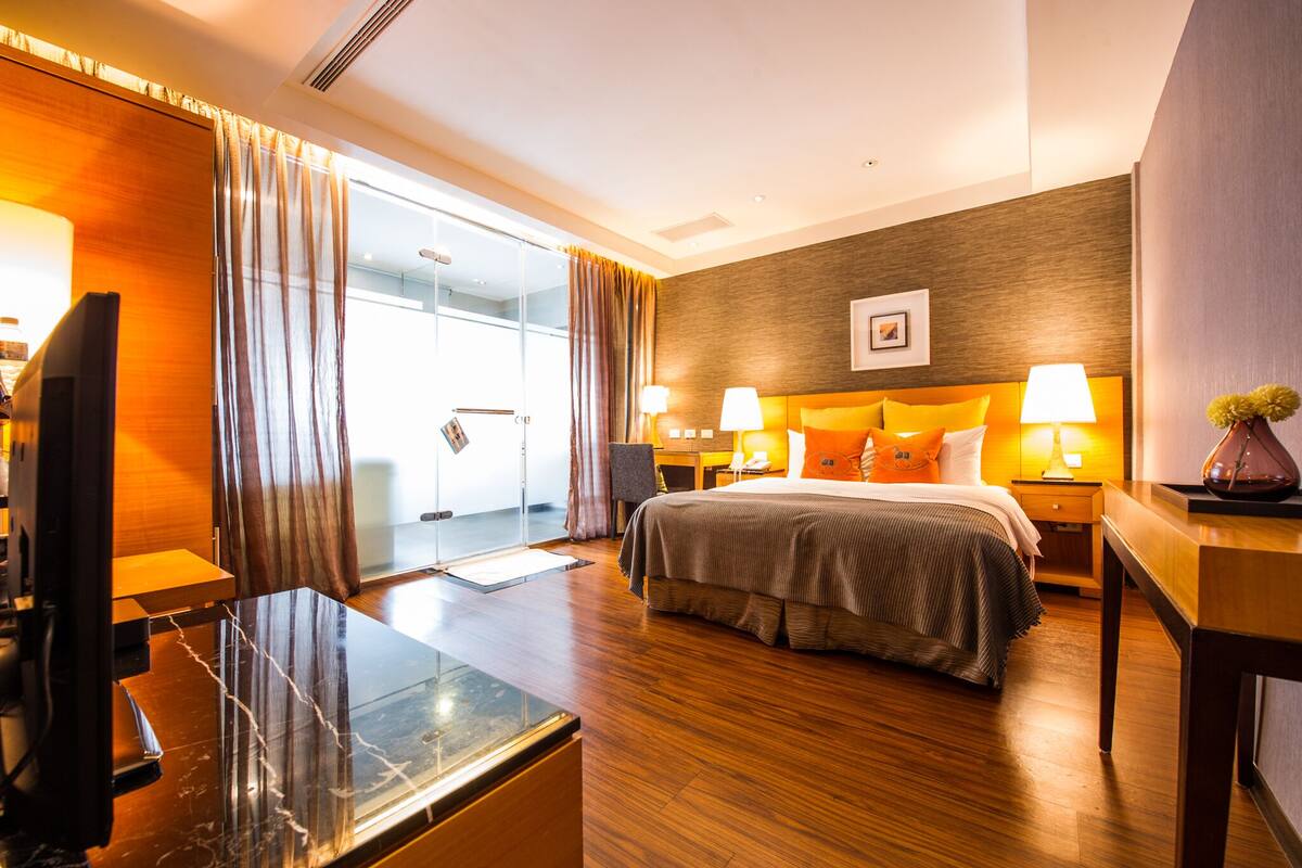 Habitación de hotel con cama, mesitas de noche, escritorio, televisor y un ventanal con cortinas.