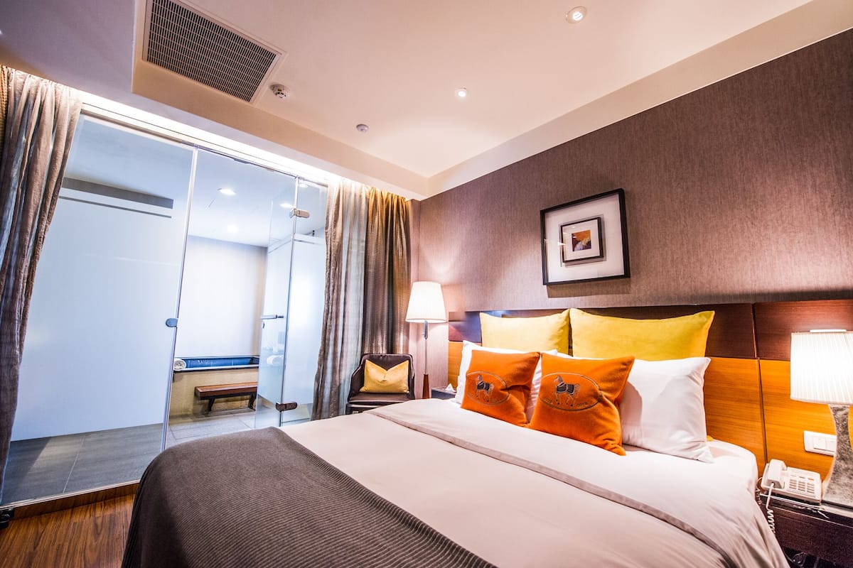 Habitación de hotel con una cama grande, un escritorio con lámpara, una silla, un banco y una puerta corredera que da a un baño.