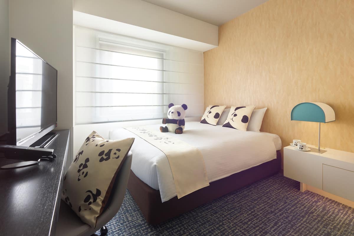 Quarto de hotel com cama, escrivaninha com televisão, um pelúcia de panda e abajur.