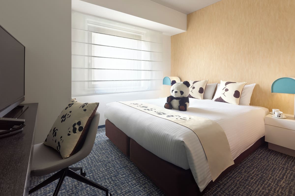 Quarto de hotel com cama, cadeira, escrivaninha, abajur e janela com persianas.