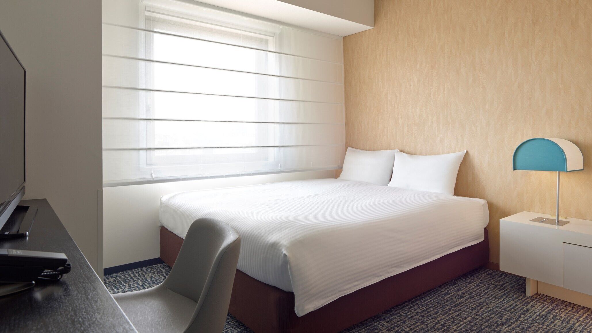 A hotel room with a bed, a desk, a chair, a lamp, and a window with blinds.