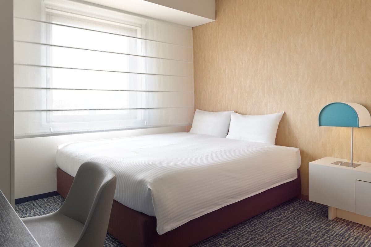 A hotel room with a bed, a desk, a chair, a lamp, and a window with blinds.