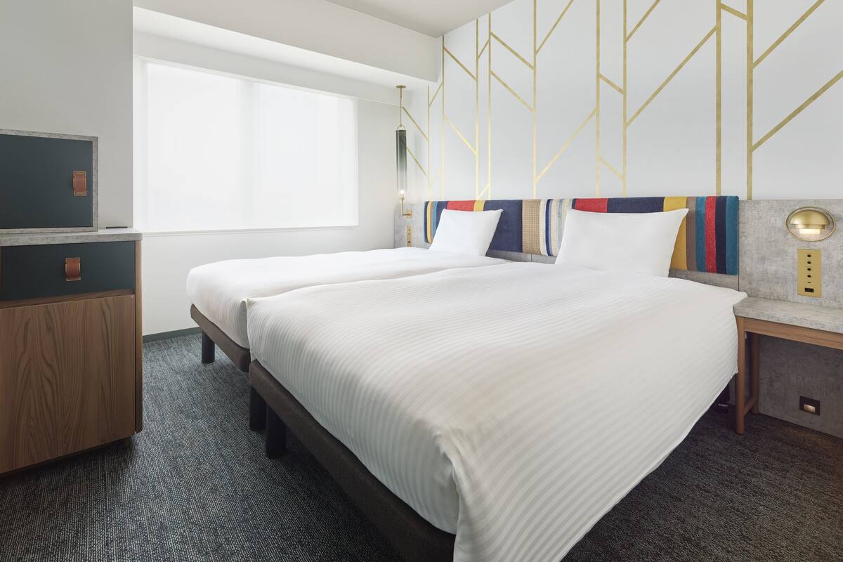 Quarto de hotel com duas camas, uma escrivaninha, uma janela grande e um design geométrico na parede.