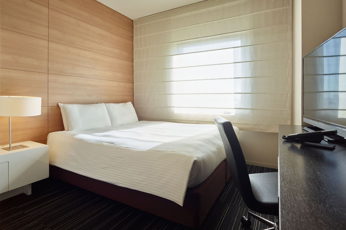 Quarto de hotel com cama, escrivaninha, cadeira, luminária e janela com persianas.