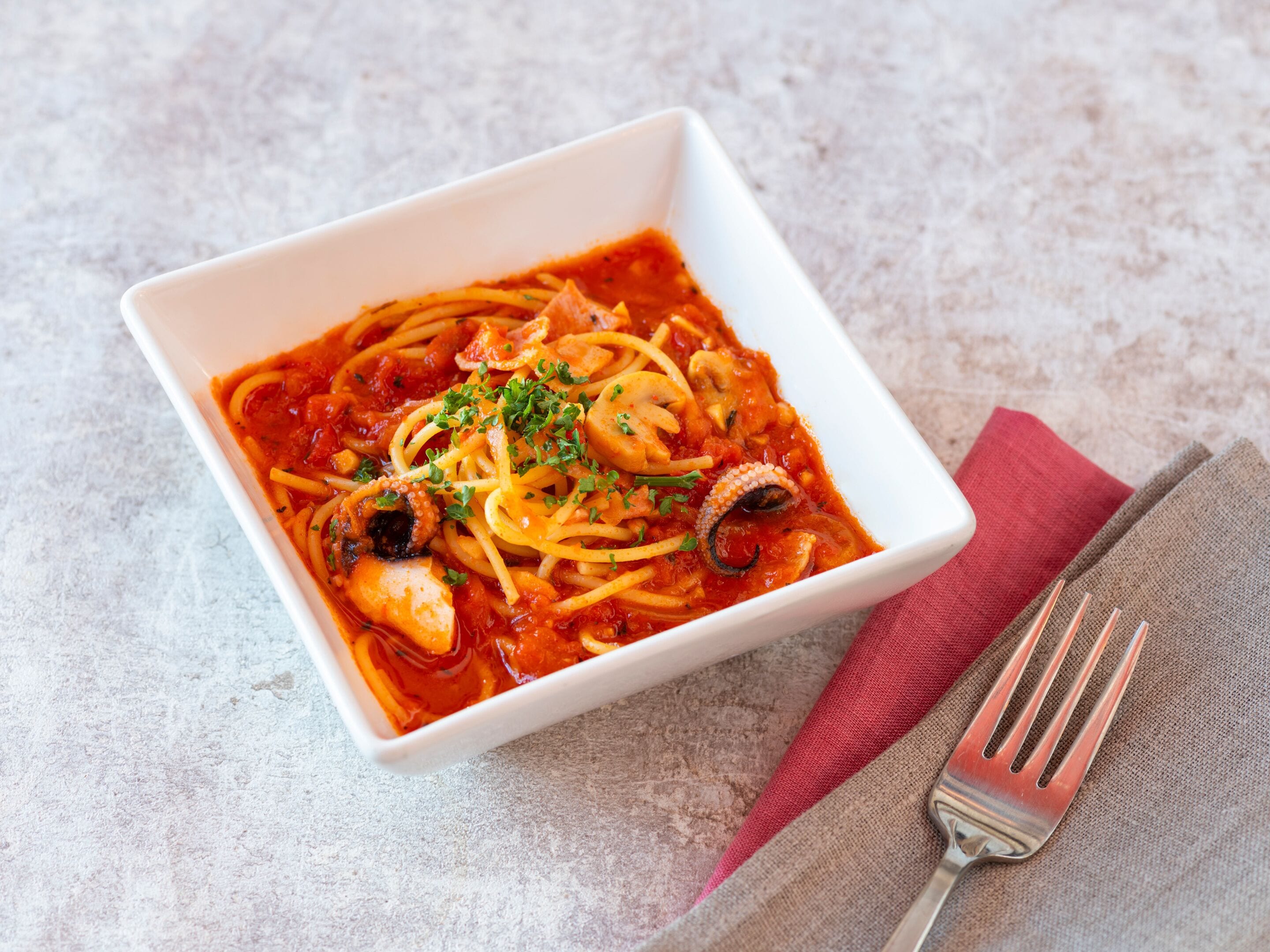 A square white bowl of spaghetti with tomato sauce, garnished with herbs, on a textured surface with a fork and napkin beside it.